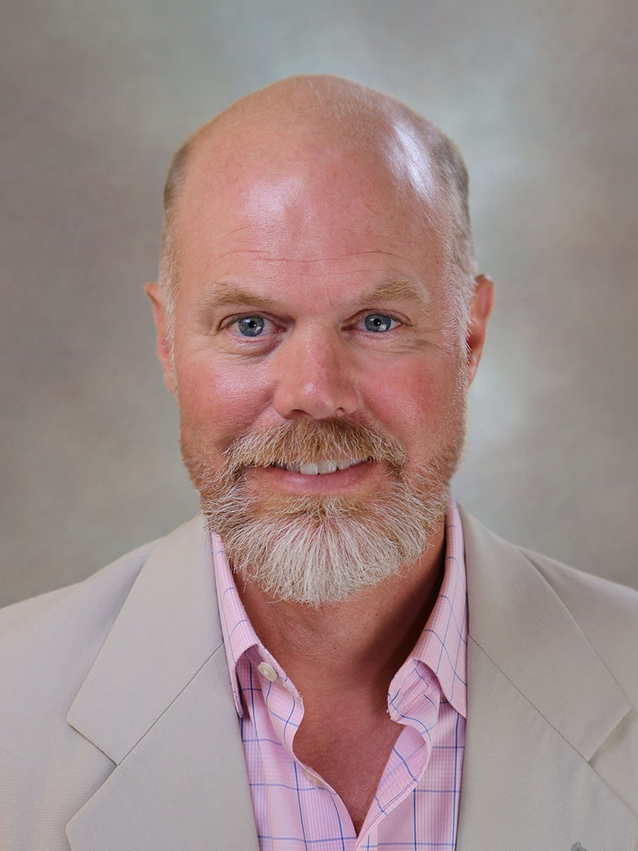 368d97582b1c94582e75d0af5120a5f1 Professional headshot of a middle-aged man with blue eyes, a goatee, wearing a light blazer and pink plaid shirt, smiling against a neutral gray background.