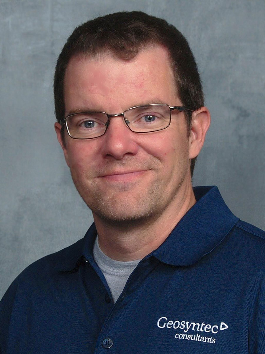 b07c4168878c5437233f490ce5adb6a4 A man wearing glasses and a blue collared shirt with the Geosyntec consultants logo smiles at the camera.
