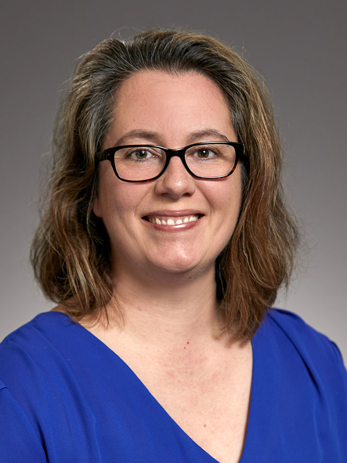 A person with shoulder-length brown hair and glasses smiles at the camera. They are wearing a bright blue top and are set against a plain, neutral background.