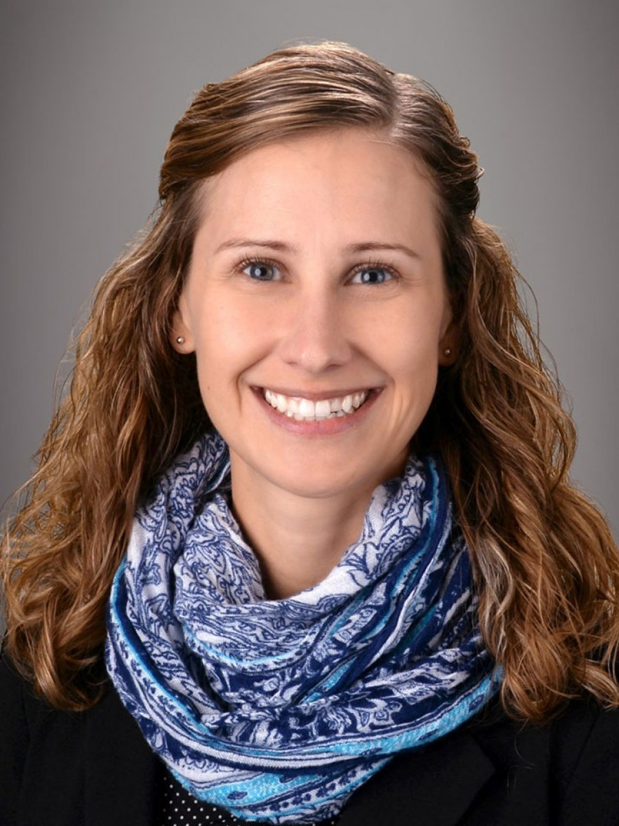A woman with wavy, light brown hair smiles at the camera. She is wearing a dark blazer and a blue and white patterned scarf, with a neutral gray background behind her.
