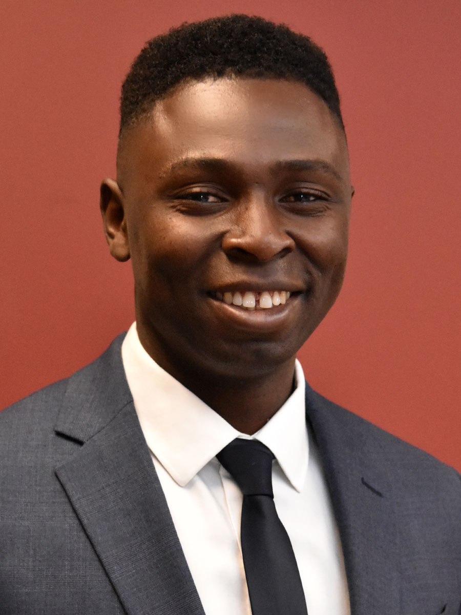 A man wearing a dark blue suit, white shirt, and black tie smiles at the camera in front of a solid reddish-brown background.