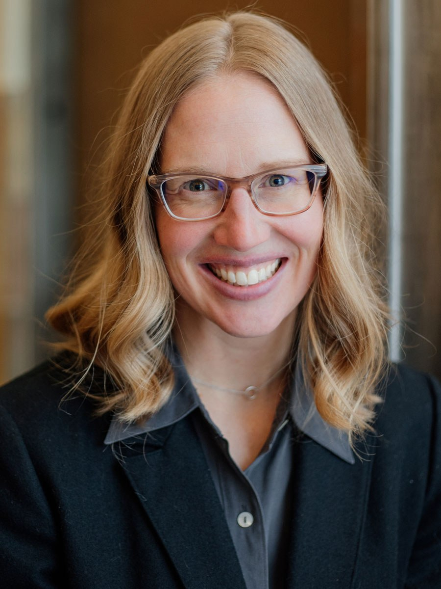 A woman with blonde, wavy hair and glasses smiles at the camera. She is wearing a dark blazer over a gray collared shirt and a delicate necklace, with a blurred indoor background.