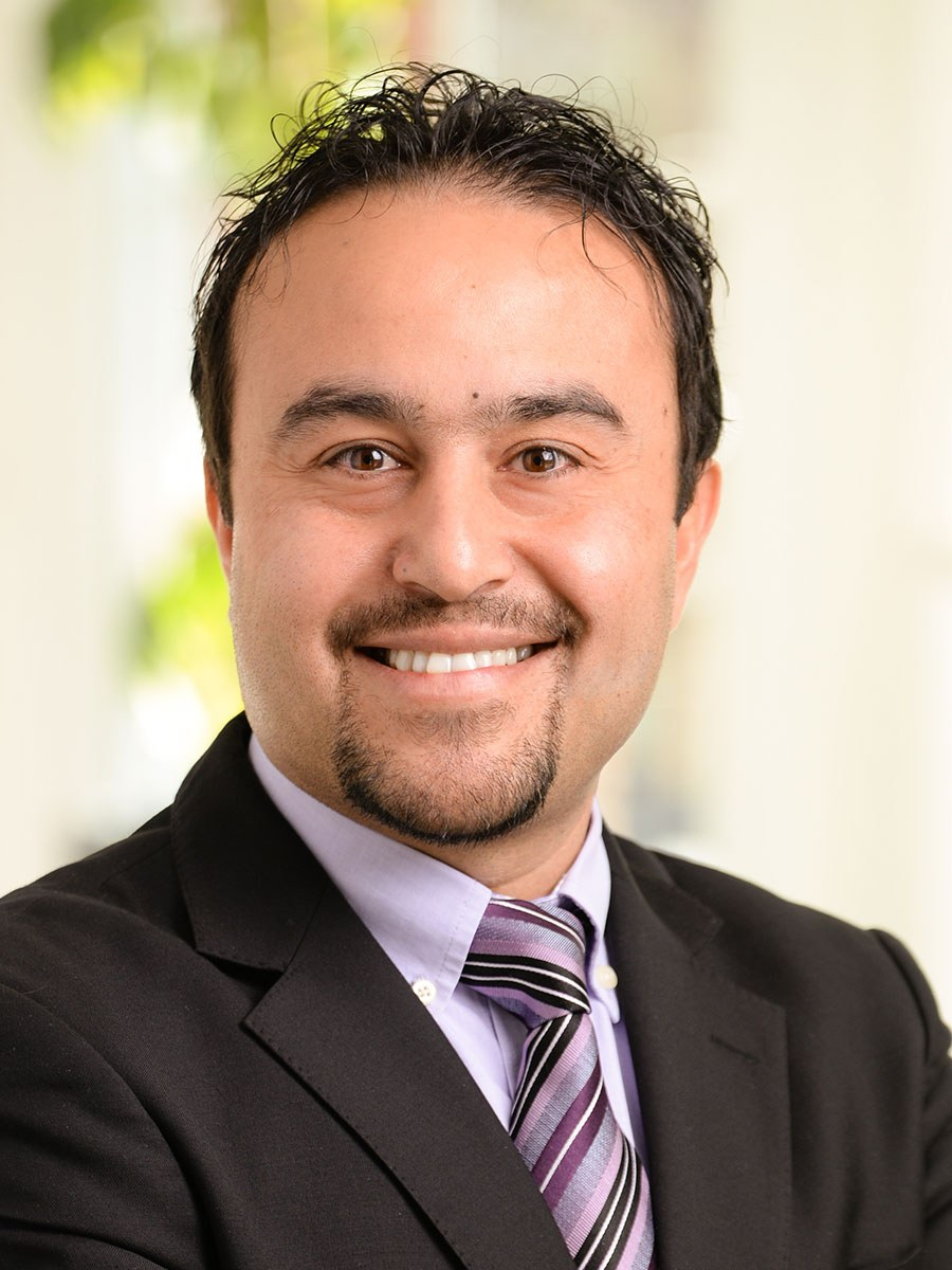 A man wearing a dark suit, light purple shirt, and striped tie smiles at the camera. He has short dark hair, a trimmed goatee, and stands in a brightly lit indoor setting with blurred green plants in the background.