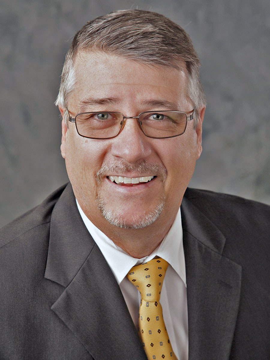 dd934f16db01c6798505dca45564caa1 A middle-aged man with short gray hair, glasses, and a goatee, smiling while wearing a dark suit, white shirt, and yellow patterned tie against a gray background.