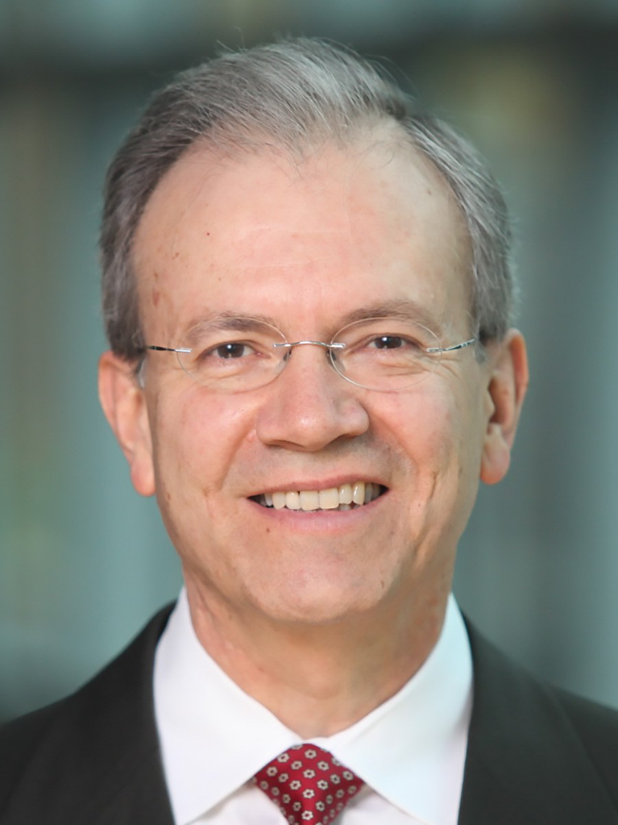 A middle-aged man with short gray hair, wearing glasses, a dark suit, white shirt, and red patterned tie, smiling at the camera with a blurred background.