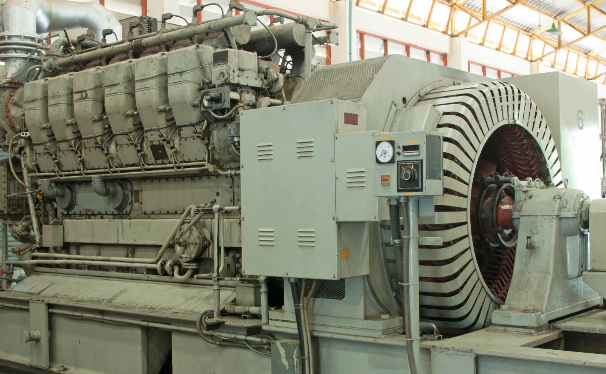 A large industrial diesel generator sits indoors, featuring a complex engine on the left and a large cylindrical generator with visible cooling fins and wiring on the right. Control panels and gauges are mounted on the side.