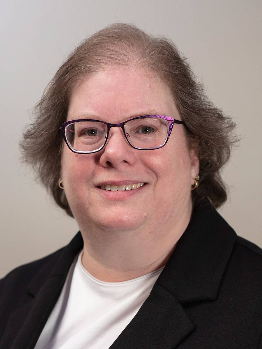 A person with short, light brown hair and glasses smiles at the camera. They are wearing a black blazer over a white shirt, with small hoop earrings, against a plain, light-colored background.