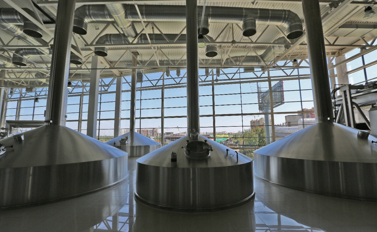Large, shiny stainless steel brewing tanks inside a modern brewery, with pipes above and large windows letting in natural light and showing an urban cityscape outside.