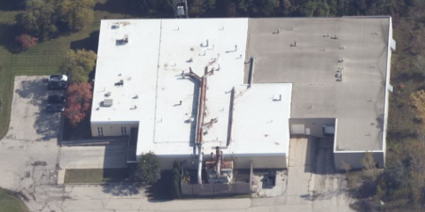 Aerial view of a large industrial building with a white roof, rooftop equipment, and surrounding trees. The building is bordered by a paved lot and grassy areas. Shadows indicate a sunny day.