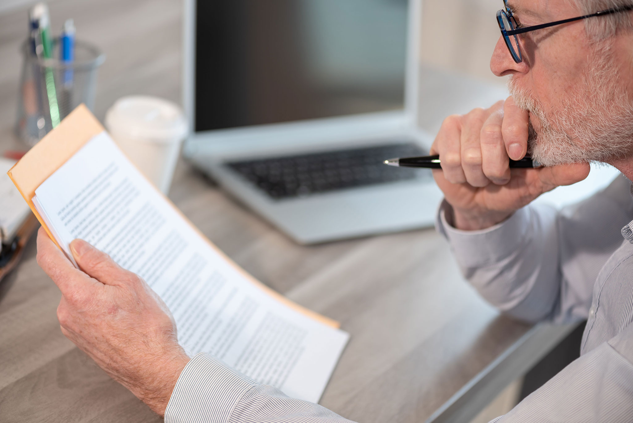 An older man with glasses sits at a desk, thoughtfully reviewing documents while holding a pen. A laptop, a coffee cup, and office supplies are on the desk in the background.