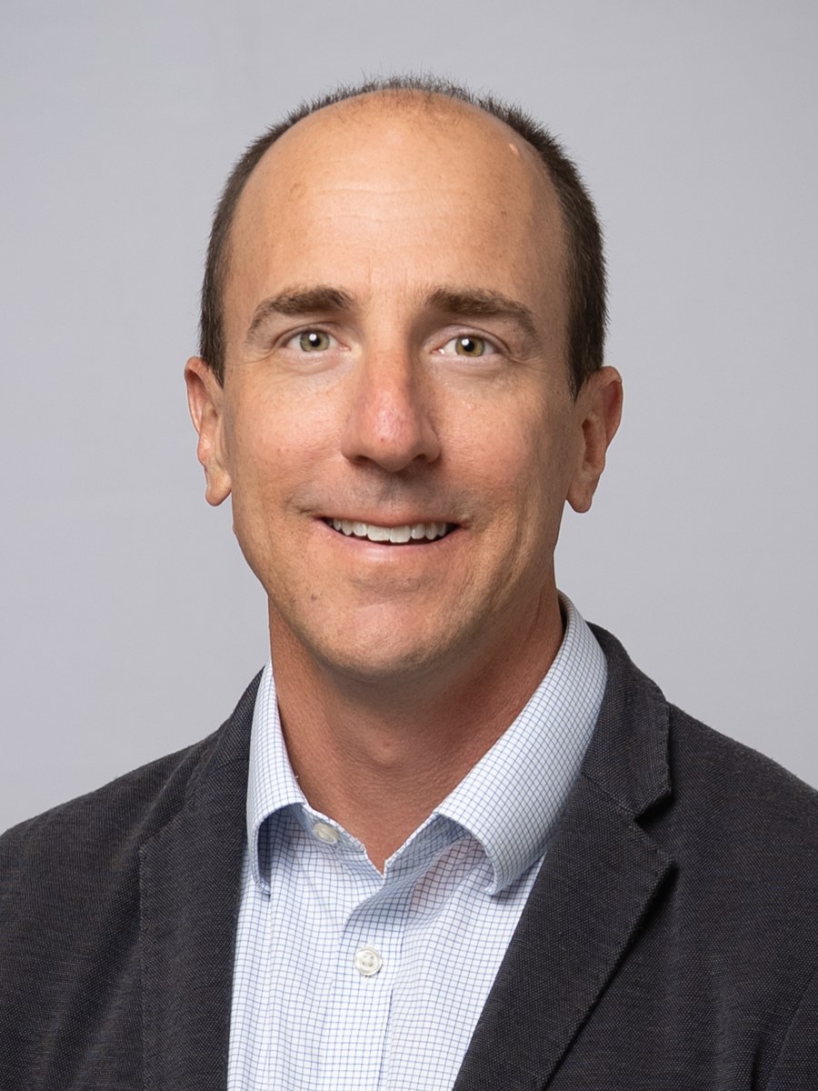 A man with short brown hair and a receding hairline smiles at the camera. He is wearing a light blue collared shirt and a dark blazer, posed against a plain light gray background.