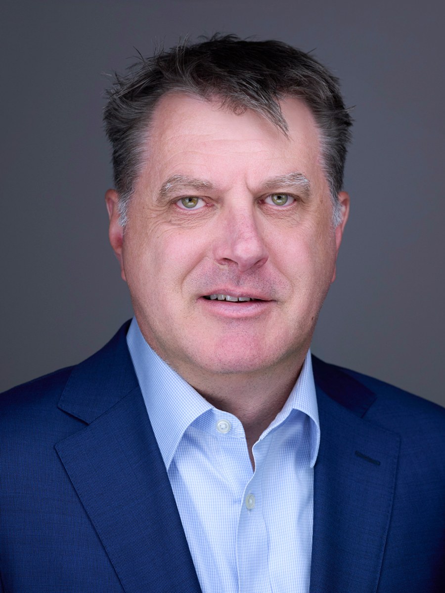 A middle-aged man with short brown hair wearing a blue suit jacket and a light blue dress shirt, posing against a plain gray background.