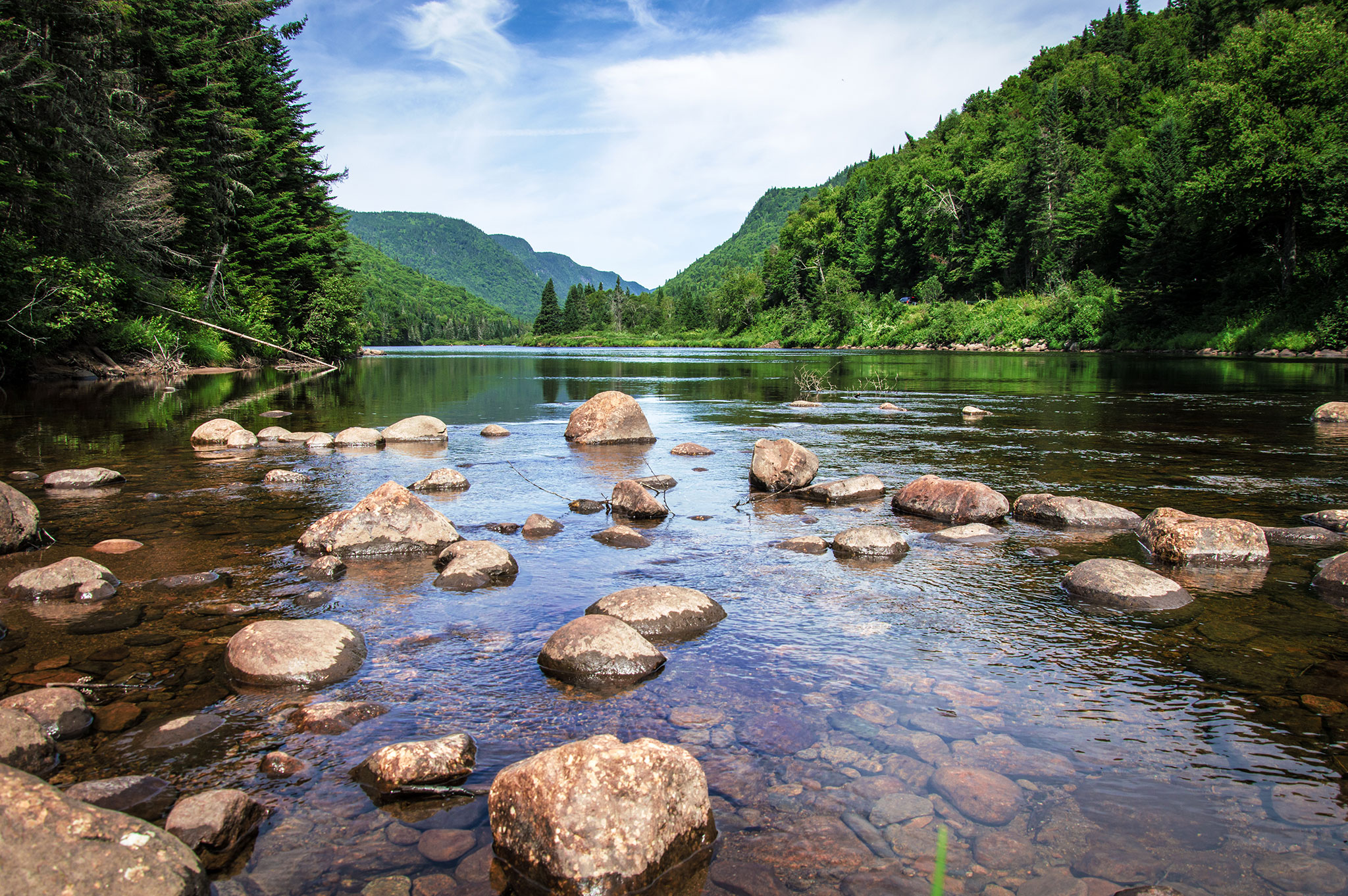 A clear river with scattered rocks flows through a green forested valley, with lush mountains and a blue sky with wispy clouds in the background.