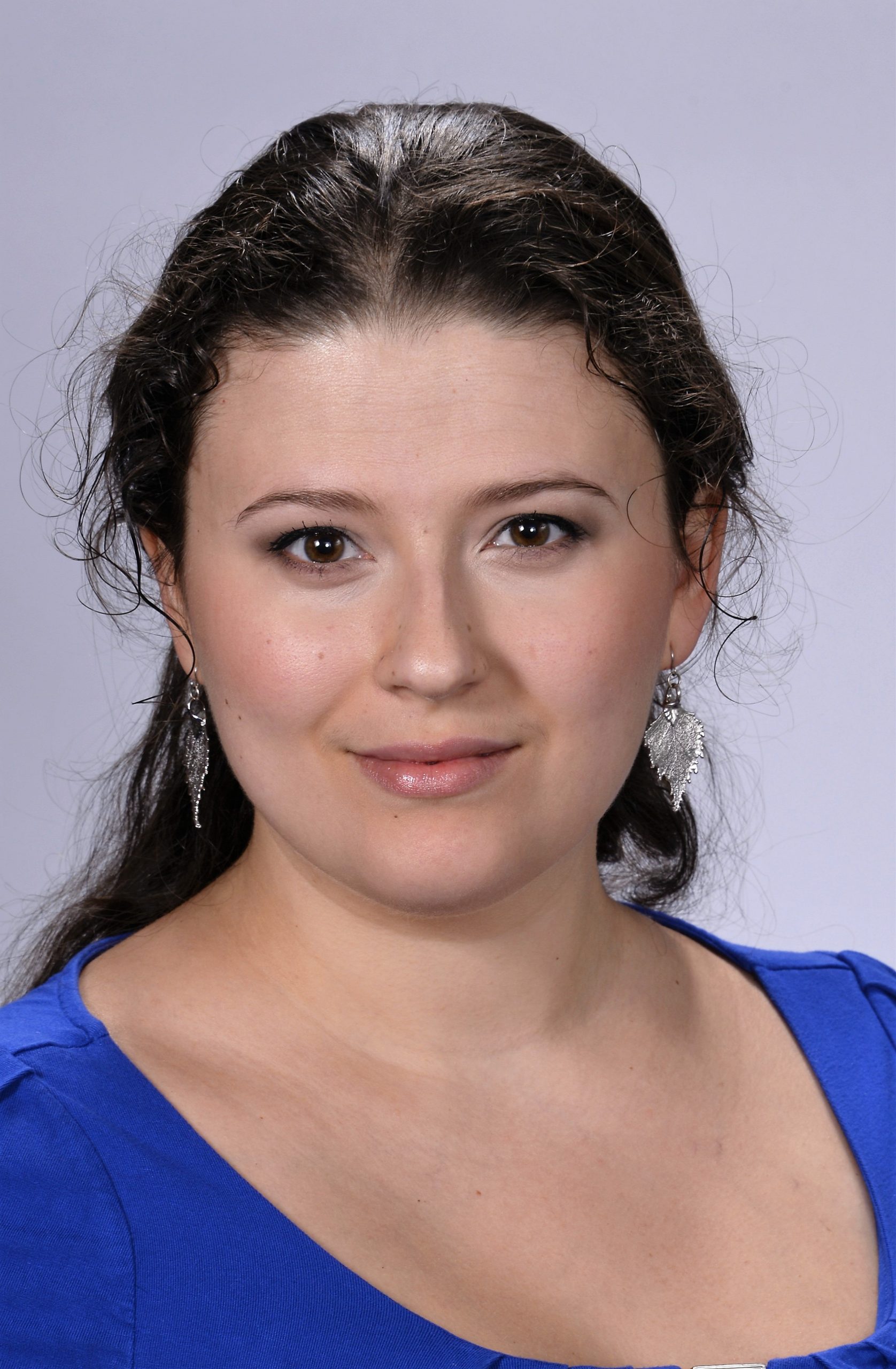 A woman with long, dark, wavy hair, wearing silver dangle earrings and a royal blue top, looks at the camera with a neutral expression against a plain light gray background.