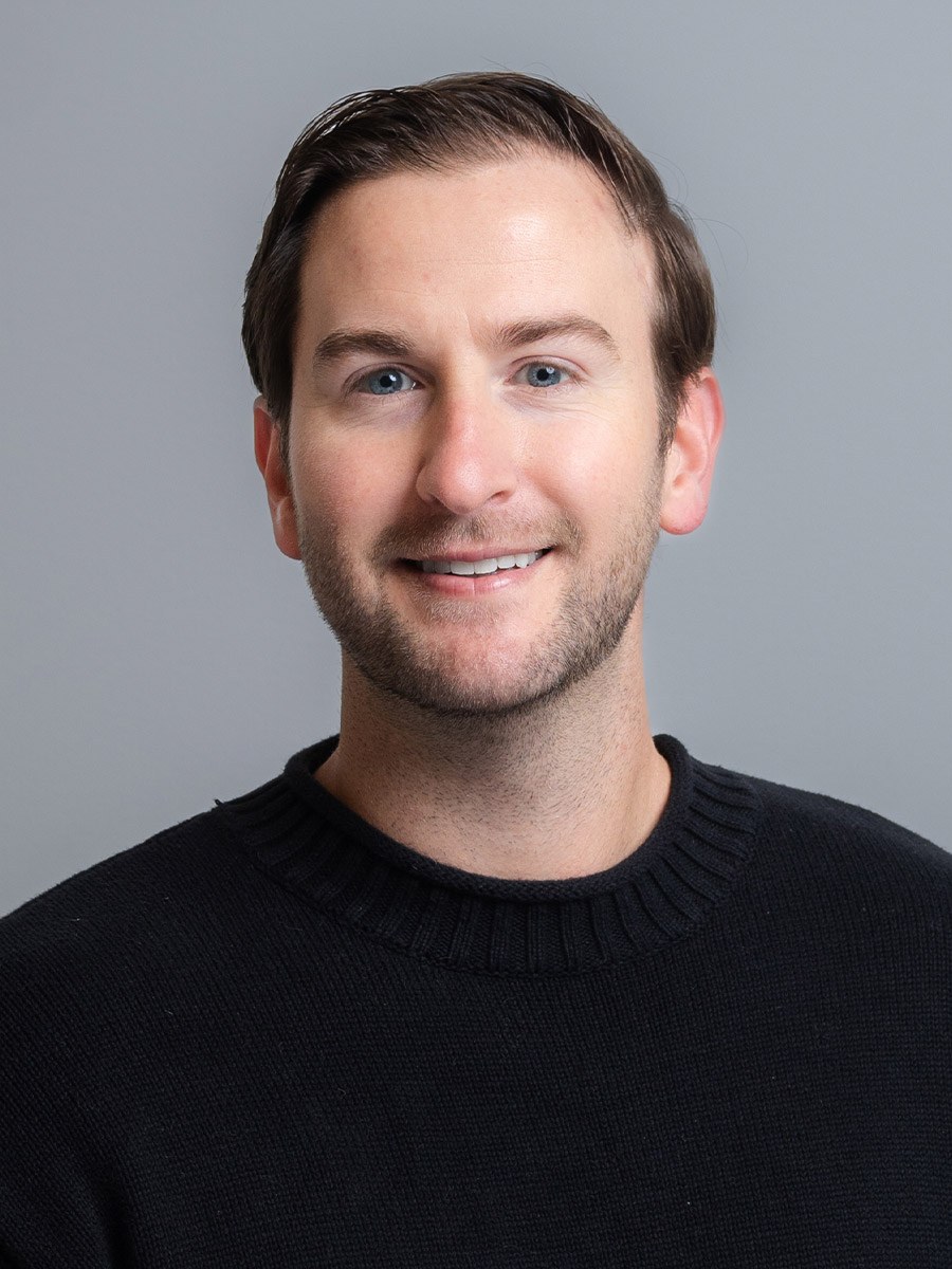 Eric Schellenger smiles for a professional headshot. He is wearing a black shirt and is in front of a dark gray background.